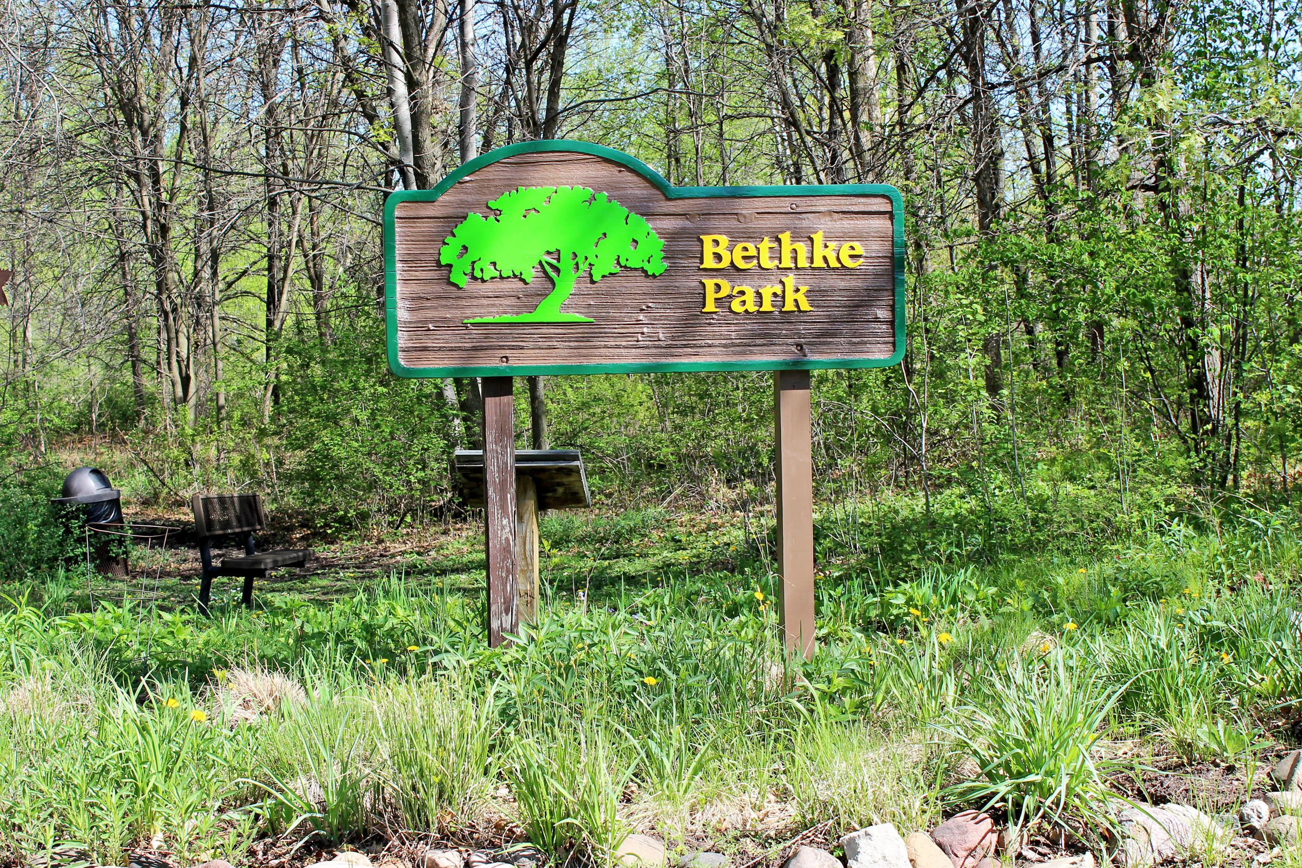 Brown sign with green tree that reads "Bethke Park" with shrubs, weeds and trees