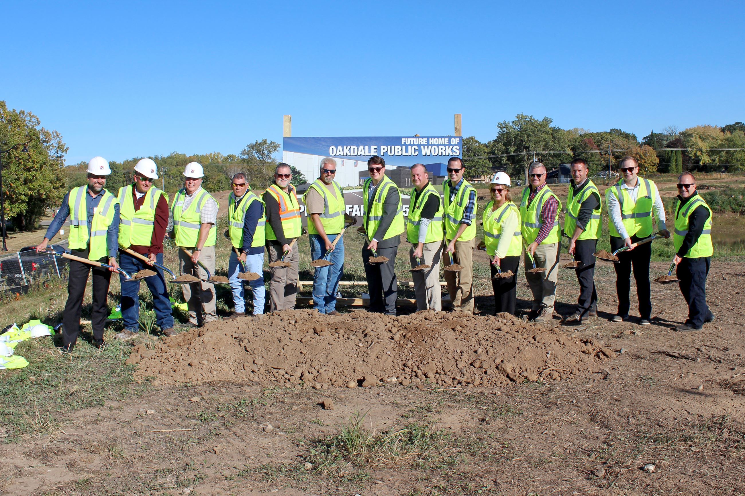 Group of 14 people in construction vests holding shovels and stand behind mound of dirt  and sign